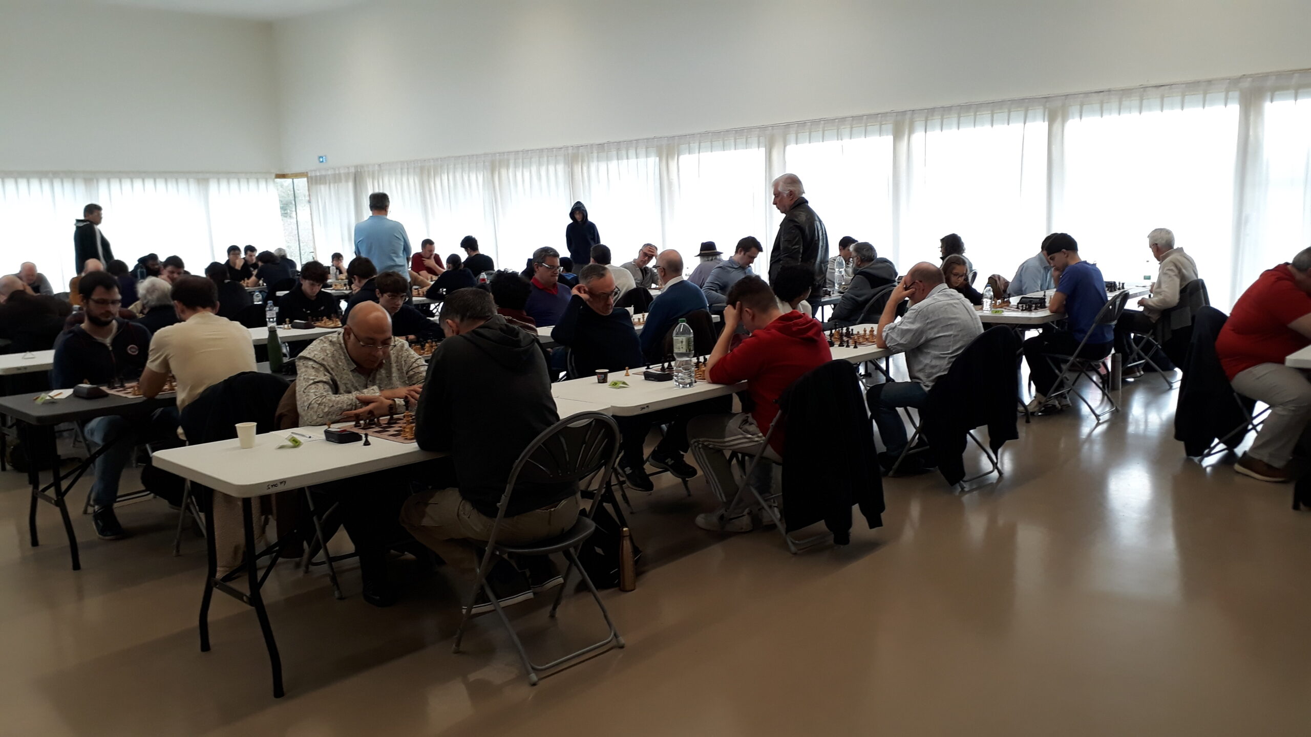 Grande salle lumineuse avec de nombreux participants jouant des parties d’échecs en tournoi.