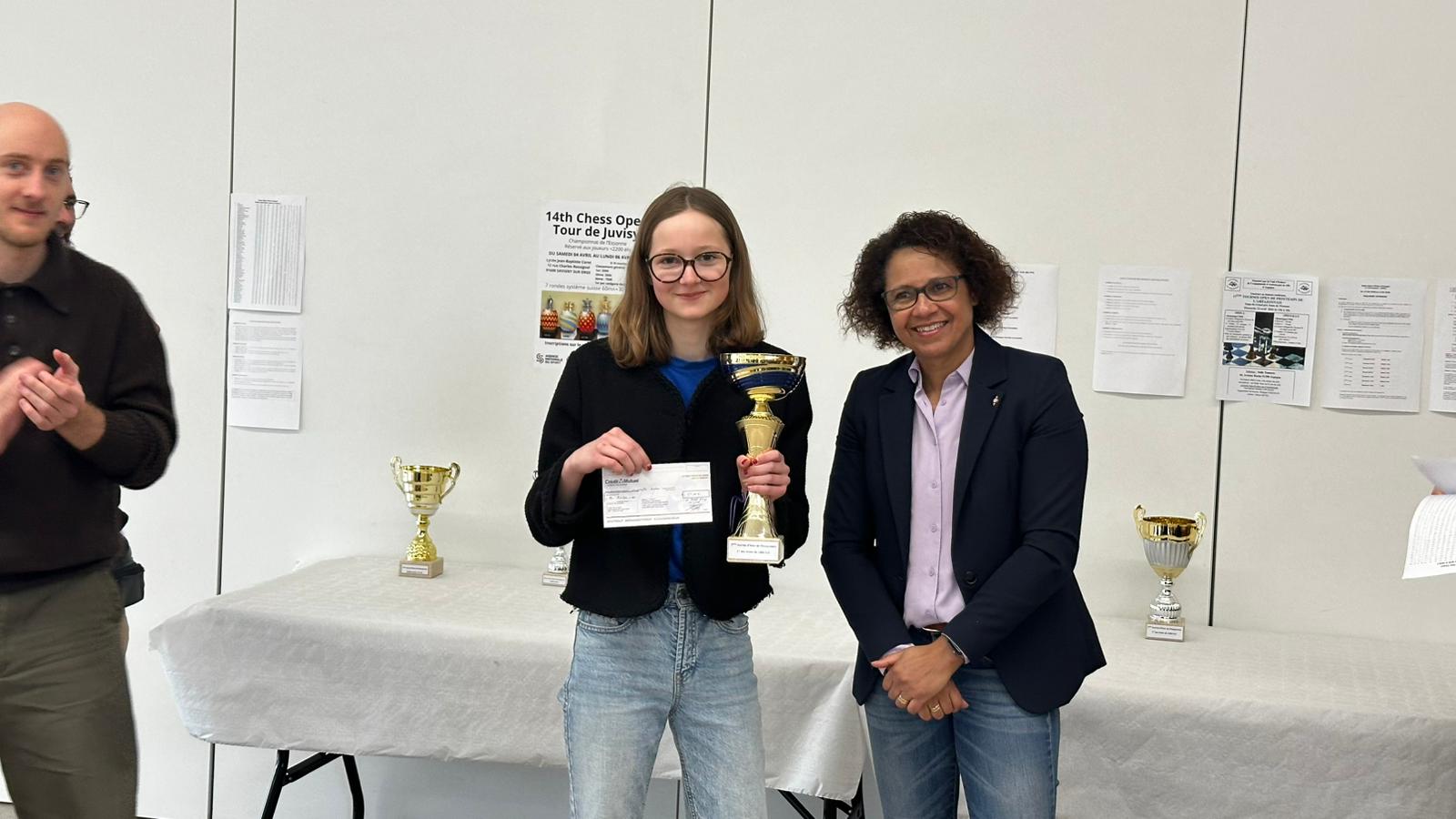 Jeune lauréate tenant une coupe rouge et or lors de la remise des prix d’un tournoi d’échecs.